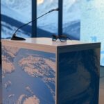 Pódio com óculos escuros em conferência de Davos, tela mostrando mapa da Groenlândia, fundo de montanhas nevadas.