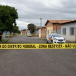 Cena de crime isolada por fita policial em rua de Santa Maria, DF, com viatura da PM ao fundo.